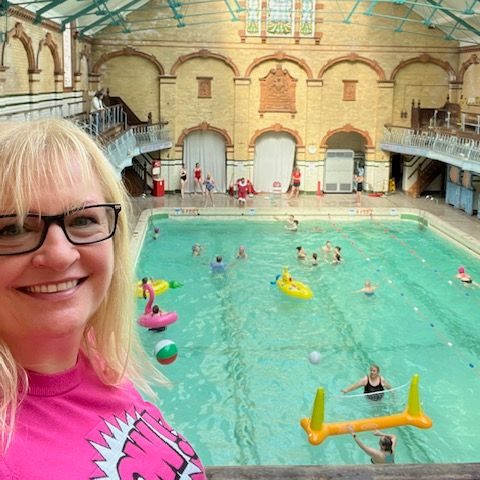 Smiling white woman with black rimmed glasses and pink jumper overlooking a heritage swimming pool filled with people and inflatables
