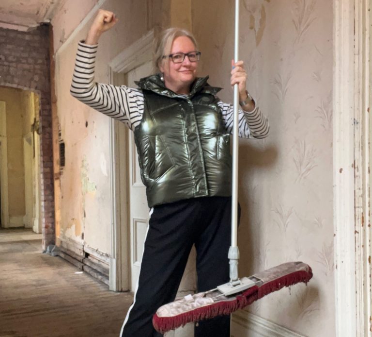 A white woman in a metallic gilet standing in a corridor in dilapidated condition holding a mop in one hand with her other arm raised 
