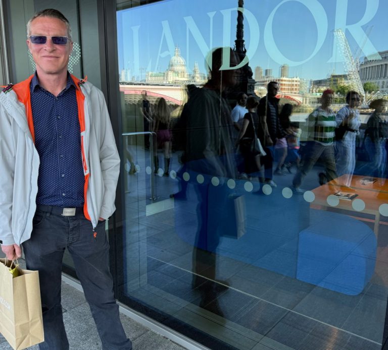 A tall white man in sunglasses standing in front of an office window showing the logo for Landor
