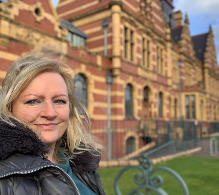 A white woman standing in front of an ornate heritage building, with terracotta and yellow tiles and scrolled metalwork